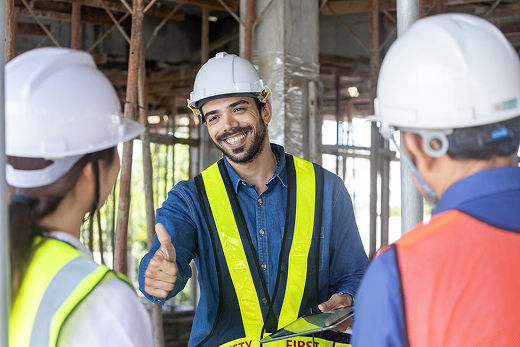 Three construction workers in white hard hats and hi-vis vests standing together on a site under scaffolding, the man in the centre smiling and giving a thumbs up
