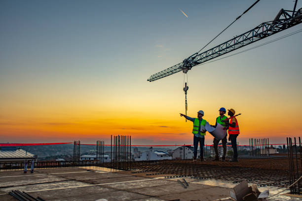 Three workers in high-visibility vests and hard hats standing on a reinforced concrete deck at sunset, one pointing up at a tower crane hook while another holds a rolled drawing