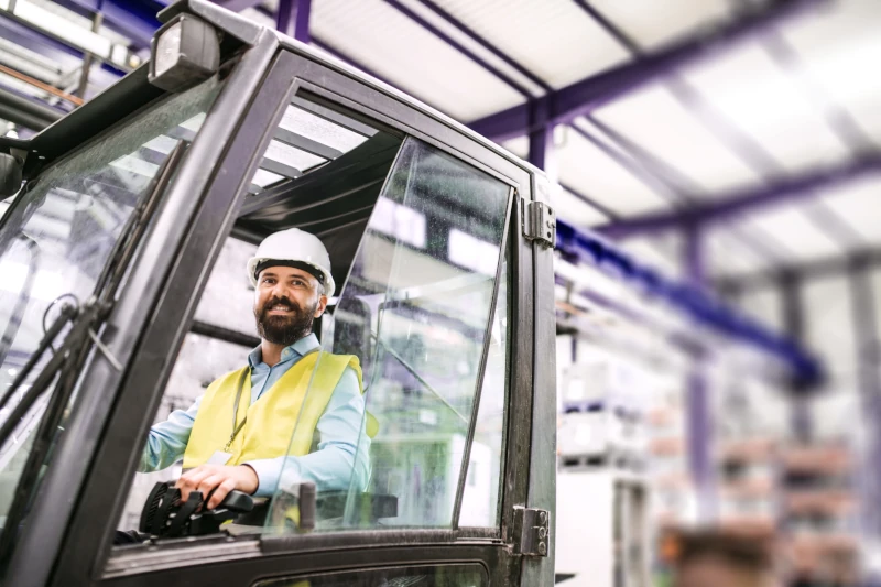 A forklift operator in a white hard hat and yellow hi-vis vest sitting in the cab of a counterbalance forklift inside a warehouse