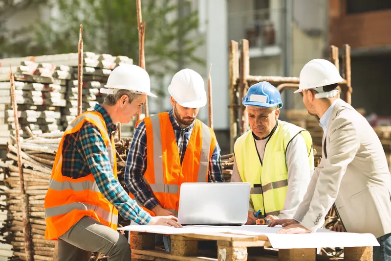 Four men in hard hats and hi-vis vests leaning over a laptop and rolled drawings on a timber pallet at a building site, with stacked formwork behind them