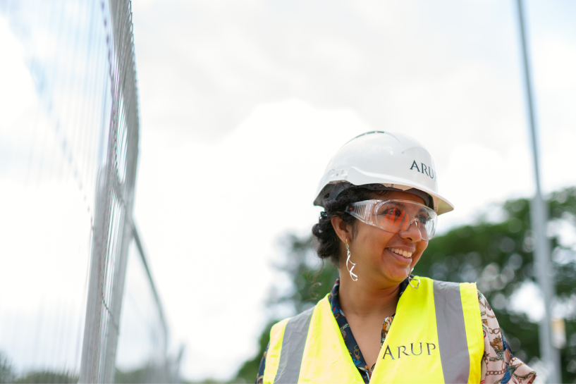 A site engineer in a white hard hat, clear safety glasses, and a yellow hi-vis vest, standing beside temporary mesh fencing on a construction site