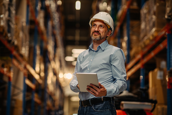 A supervisor in a white hard hat and blue shirt holding a tablet, standing in the aisle of a warehouse between tall racking bays stacked with pallets