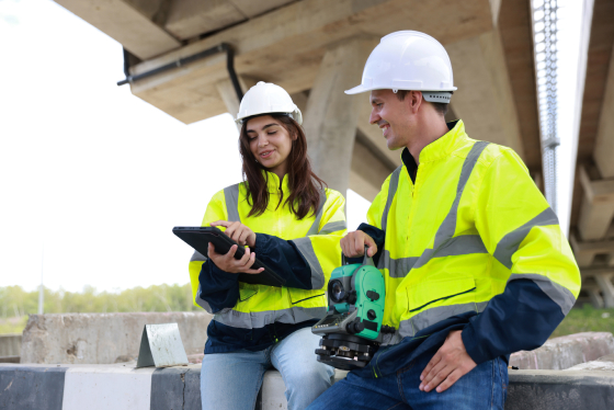 Two workers in hi-vis jackets and white hard hats sitting on a concrete barrier under a bridge, one holding a tablet, the other standing beside a surveying instrument on a tripod