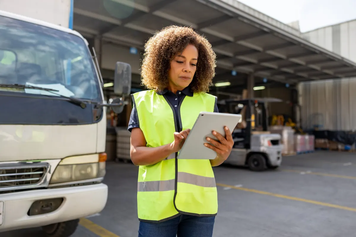 A woman in a hi-vis vest using a tablet beside the cab of a small truck at a covered loading dock, with a forklift and stacked pallets behind her