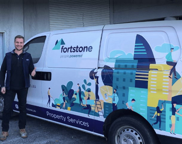 A man in a dark jacket giving a thumbs up beside a white Fortstone Property Services van wrapped in illustrated graphics of trades and buildings