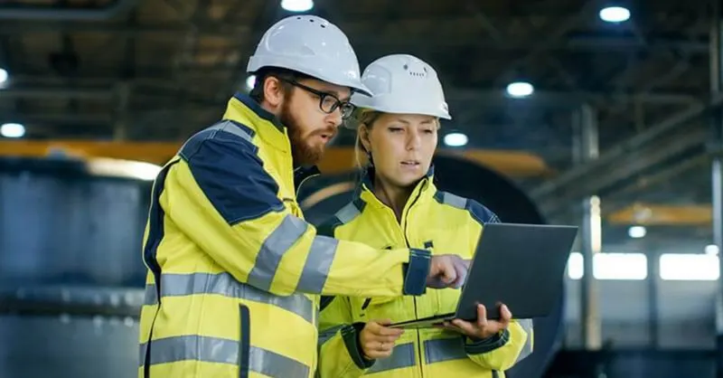 Two workers in yellow hi-vis jackets and white hard hats inside a large industrial shed, looking together at an open laptop held by one of them