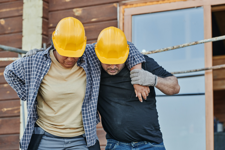 A worker in a yellow hard hat supporting an injured colleague, also in a hard hat, as they walk away from a timber-framed building under construction