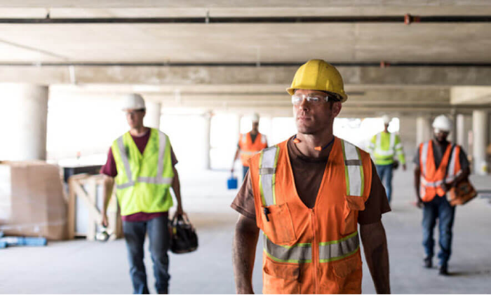 Five workers in high-visibility vests and hard hats walking across the bare concrete floor of a building under construction, one of them carrying a tool bag