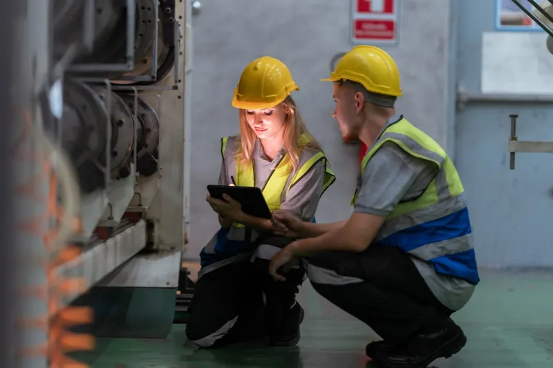 Two workers in yellow hard hats and hi-vis vests crouched beside a large industrial machine in a plant room, one writing on a tablet with a stylus