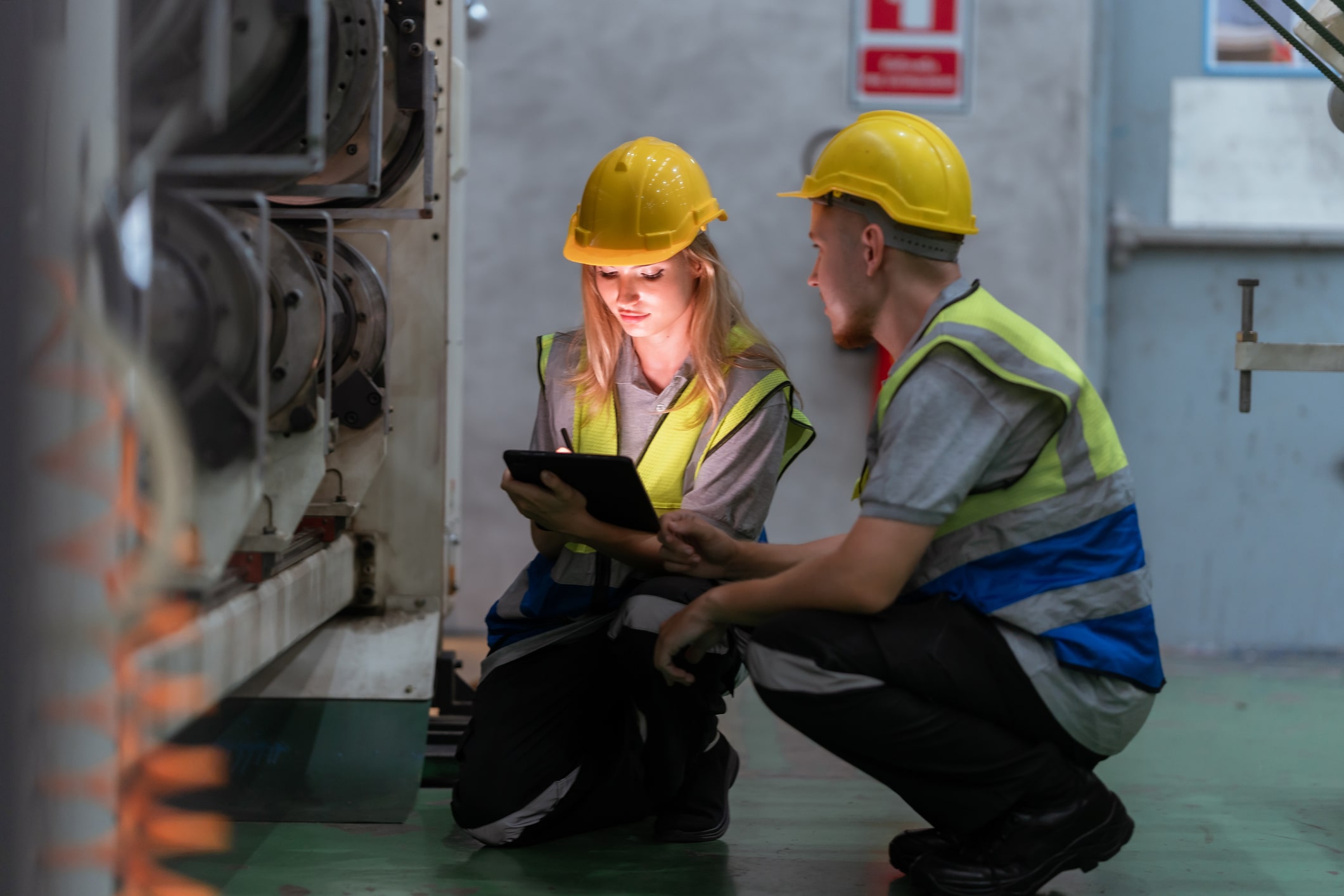 Two workers in yellow hard hats and hi-vis vests crouched beside a large industrial machine in a plant room, one writing on a tablet with a stylus