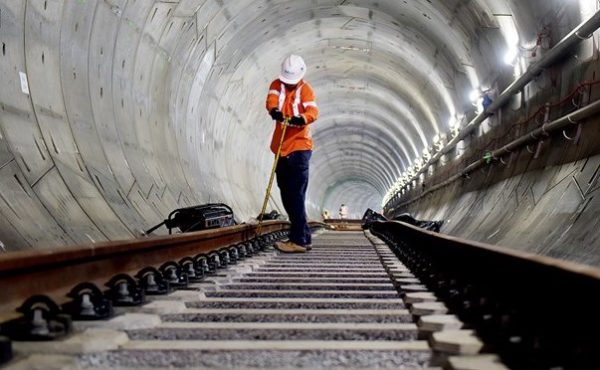 A worker in an orange high-visibility jacket and white hard hat stands on newly laid railway track inside a concrete-segment tunnel, with tools on the sleepers and more workers further along the bore