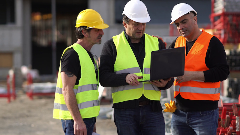 Three workers in high-visibility vests and hard hats on a construction site, two of them reading a laptop screen held open by the man in the middle