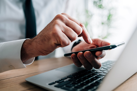 Close-up of a man in a white shirt and dark tie tapping a mobile phone screen with one hand while resting the other on an open laptop keyboard