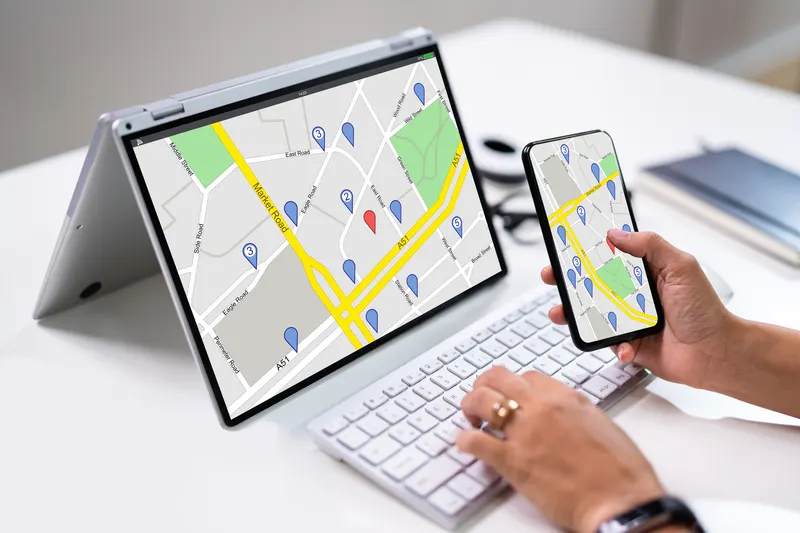 A hand holding a phone beside an open convertible laptop on a white desk, both screens showing the same street map with numbered blue location pins and one red one