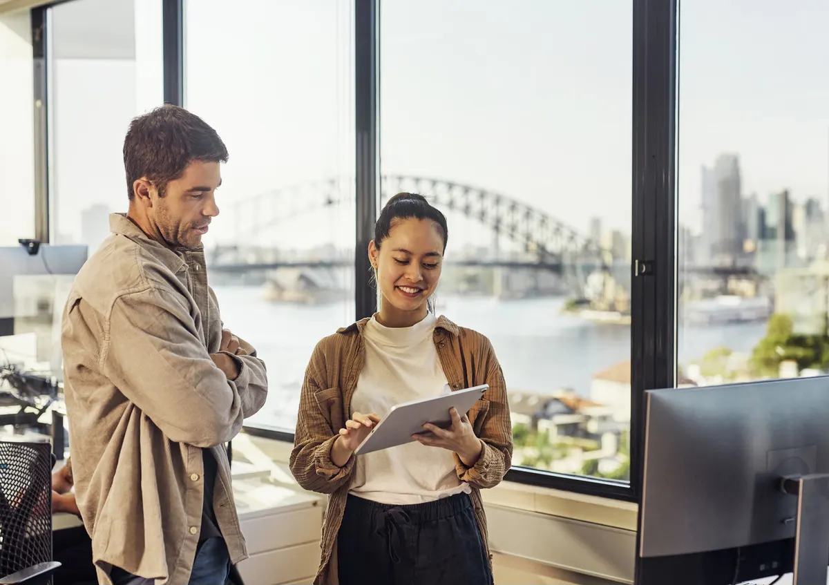 A man with folded arms and a woman holding a tablet talking beside an office window, the Sydney Harbour Bridge and harbour visible behind them