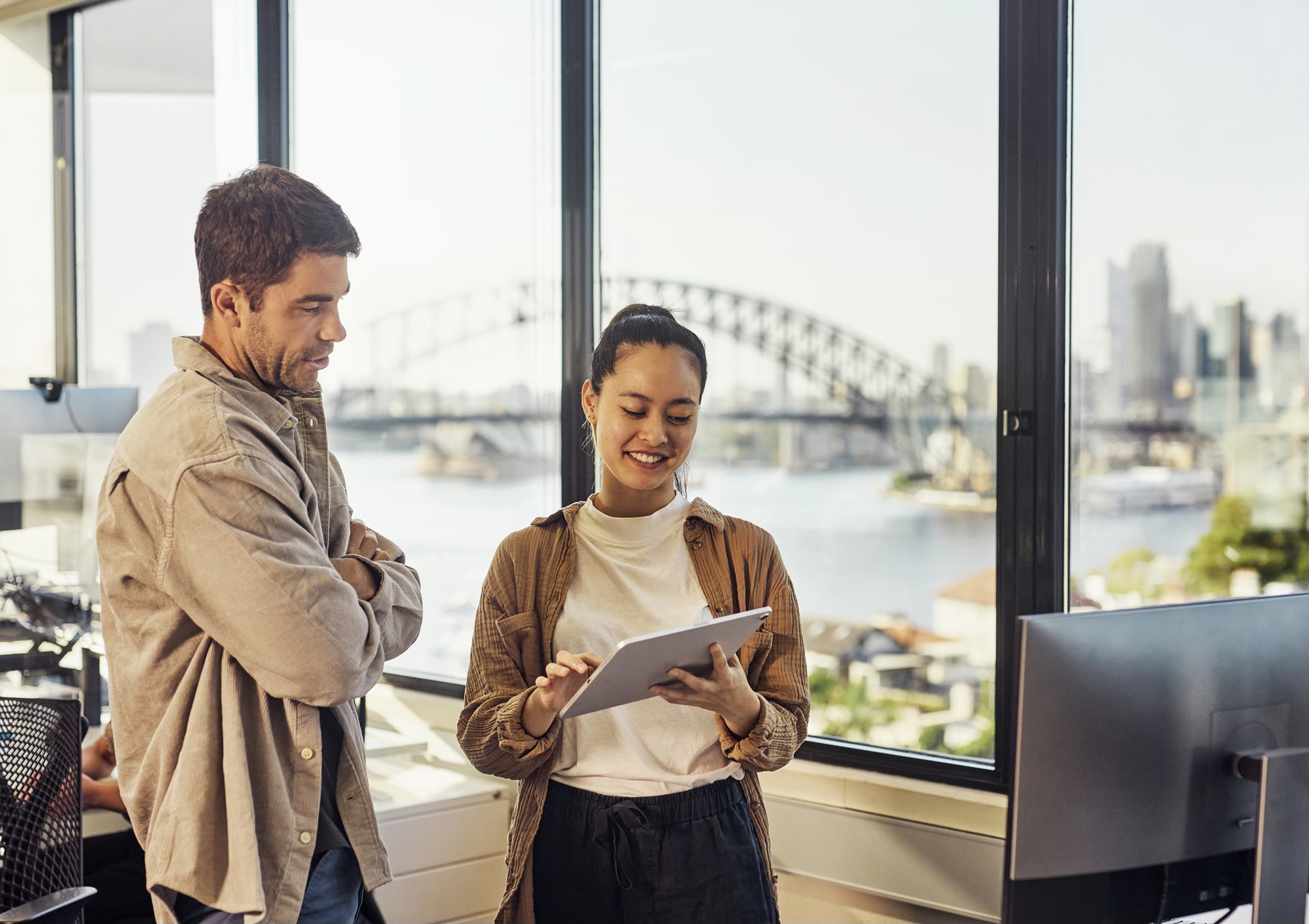 A man with folded arms and a woman holding a tablet talking beside an office window, the Sydney Harbour Bridge and harbour visible behind them