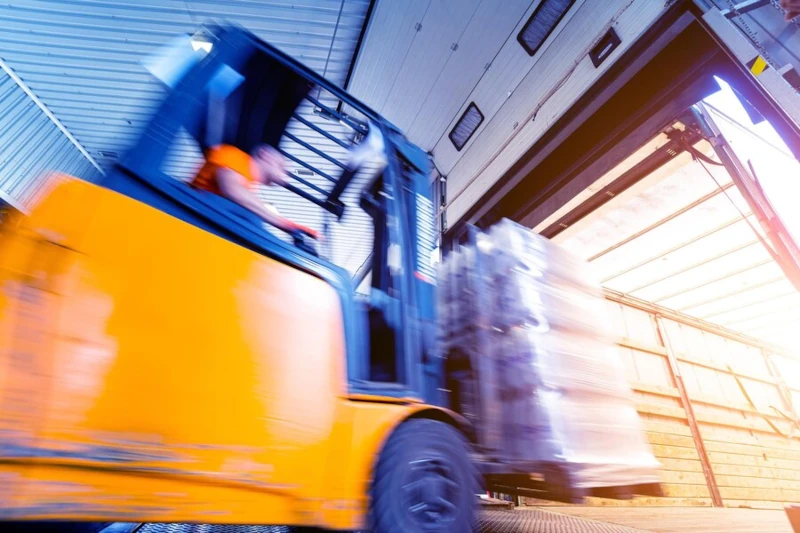 A forklift lifting a shrink-wrapped pallet into a shipping container at a warehouse loading dock, the shot blurred by the machine's motion