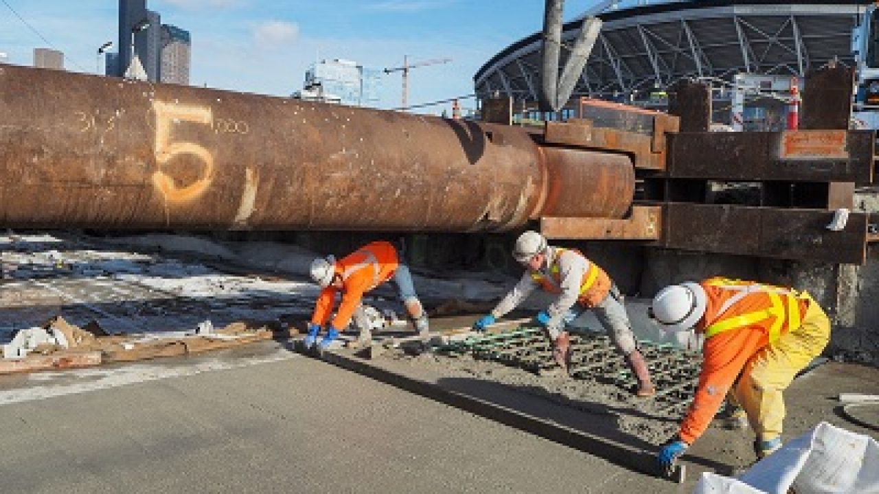 Three workers in hi-vis and hard hats screeding wet concrete over steel reinforcing mesh, under a large rusted steel pipe on temporary supports at a city construction site