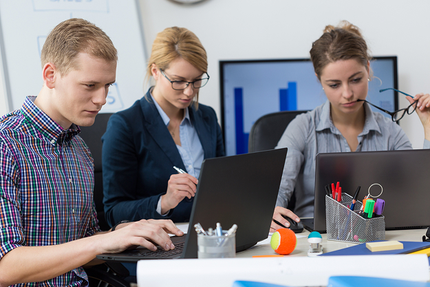 Three colleagues working at laptops around one desk in a bright office, with a bar chart on the wall screen behind them and pens and notes spread across the table