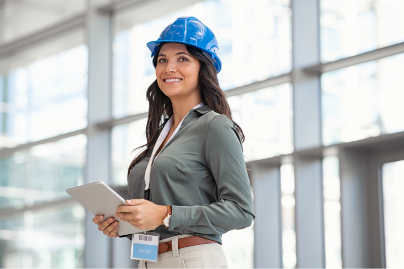 A woman in a blue hard hat with a visitor pass clipped to her belt, holding a tablet in the bright glass-walled lobby of an office building