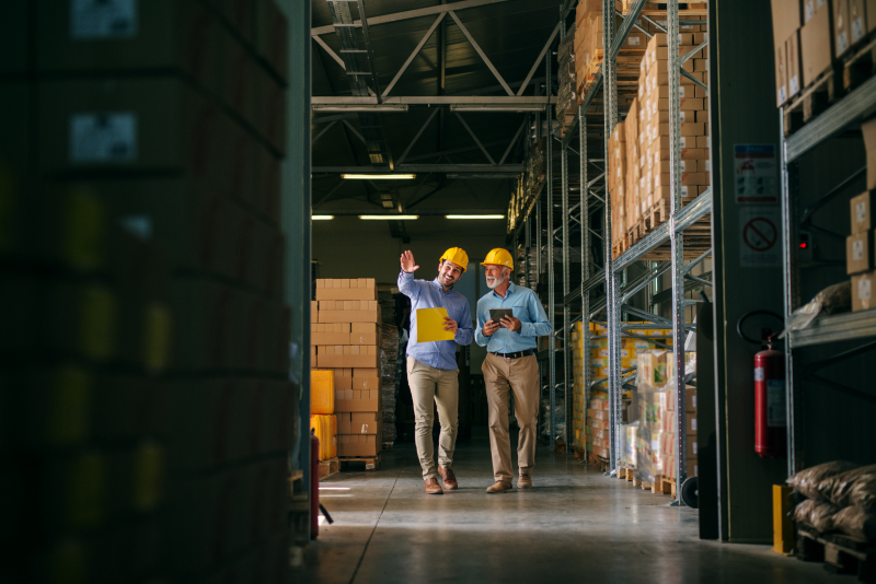 Two men in yellow hard hats walking down an aisle of a warehouse between racked pallets, one carrying a yellow clipboard and the other a tablet